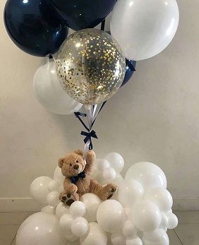 Brown 12 inches Teddy bear sitting on 20 white small and large blown balloons holding a stick of 1 Clear gold confetti balloon and 5 Black and white balloons
