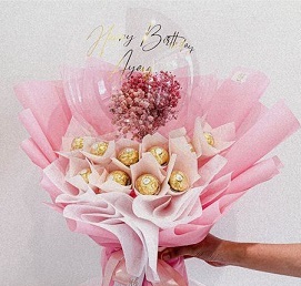 Transparent Balloon with print message happy Birthday and stuffed with small flowers tied on the outside are 16 Ferrero rocher chocolates wrapped in pink and white
