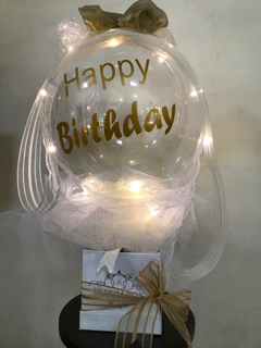 Happy birthday print on the transparent balloon white net wrapping and gold bow in a box with LED string lights