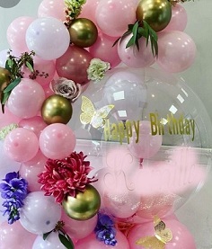 Happy birthday print on the transparent stuffed balloon with small and large light pink white and golden balloons garland decorated with butterflies and red and white flowers