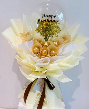 Transparent Balloon with print message happy Birthday and stuffed with small flowers tied on the outside are 16 Ferrero rocher chocolates wrapped in cream and white